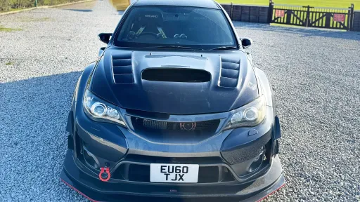 Front view of black Subaru Impreza with bonnet scoop and sporty grille, parked on gravel with open countryside in the background.