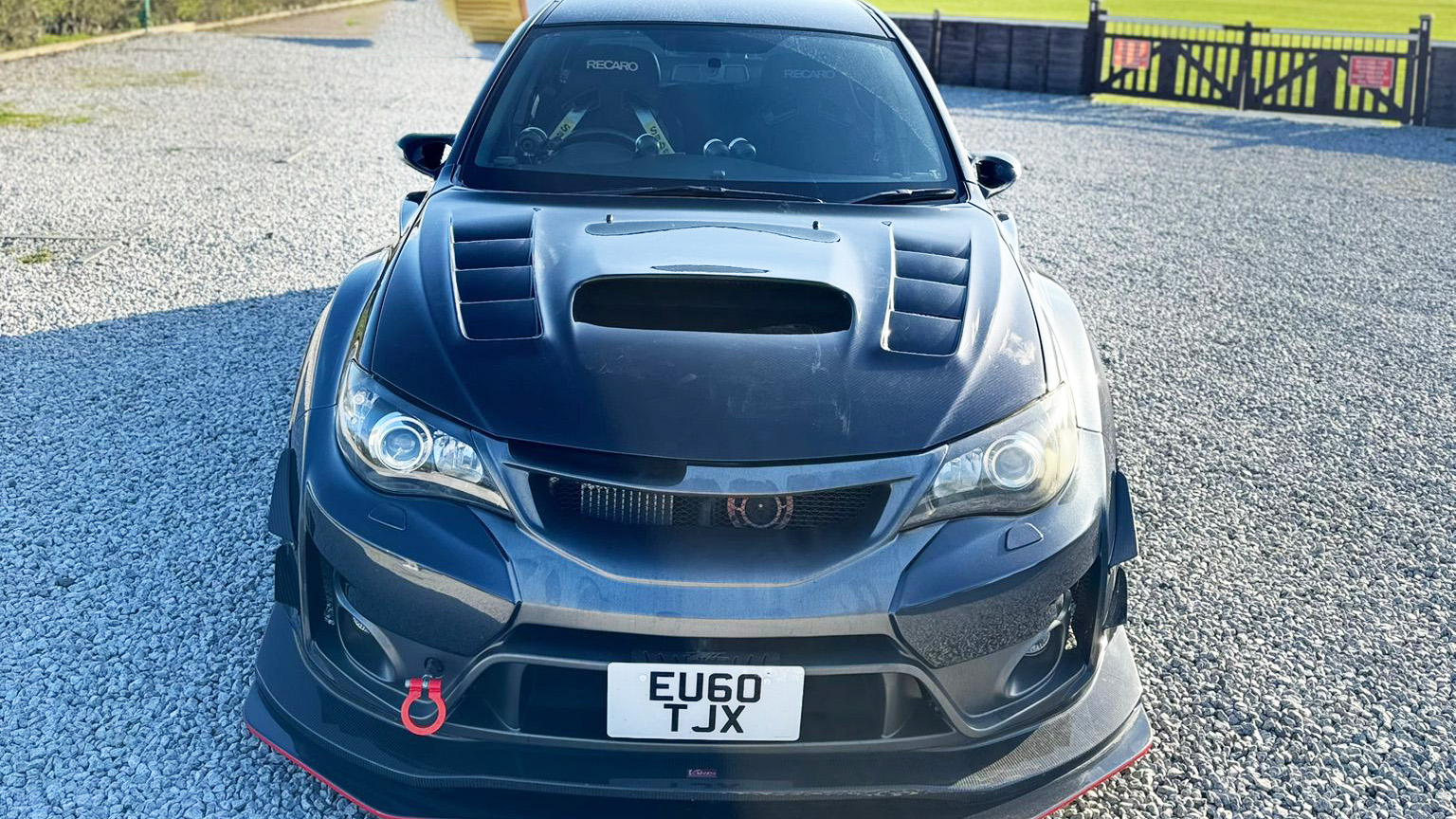 Front view of black Subaru Impreza with bonnet scoop and sporty grille, parked on gravel with open countryside in the background.