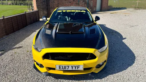 Front view of yellow Ford Mustang GT with black carbon bonnet and aggressive grille, parked on gravel with countryside background.