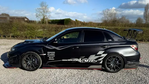 Side profile of black Subaru Impreza with racing decals and alloy wheels, parked on gravel with trees and countryside behind.