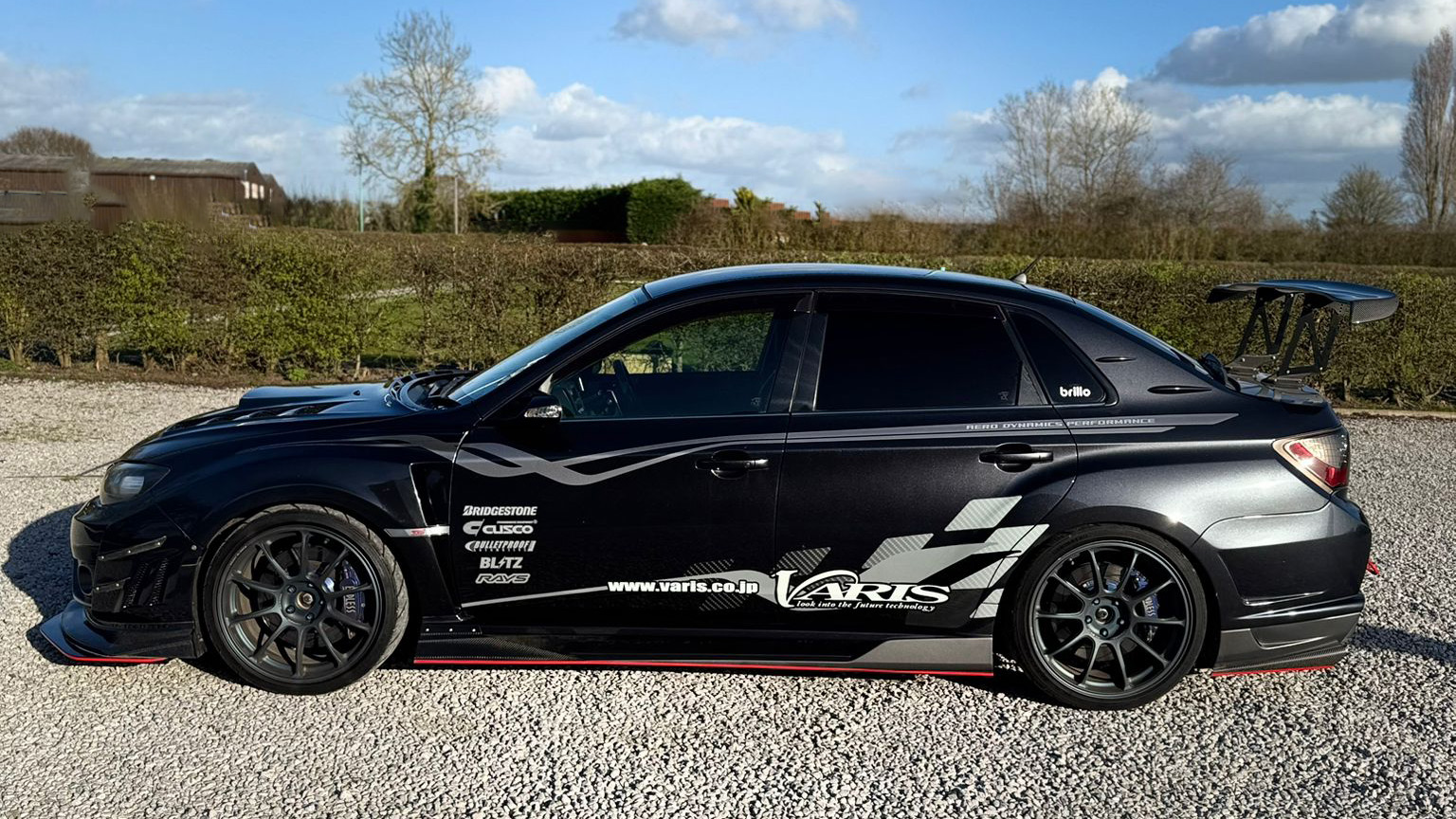 Side profile of black Subaru Impreza with racing decals and alloy wheels, parked on gravel with trees and countryside behind.