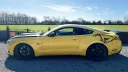 Side profile of yellow Ford Mustang GT with black roof and running horse graphics, parked on gravel with open fields behind.