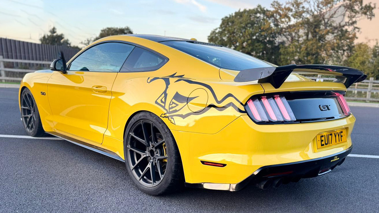 Rear three-quarter view of yellow Ford Mustang GT with black roof, rear spoiler and black wheels, parked on gravel.