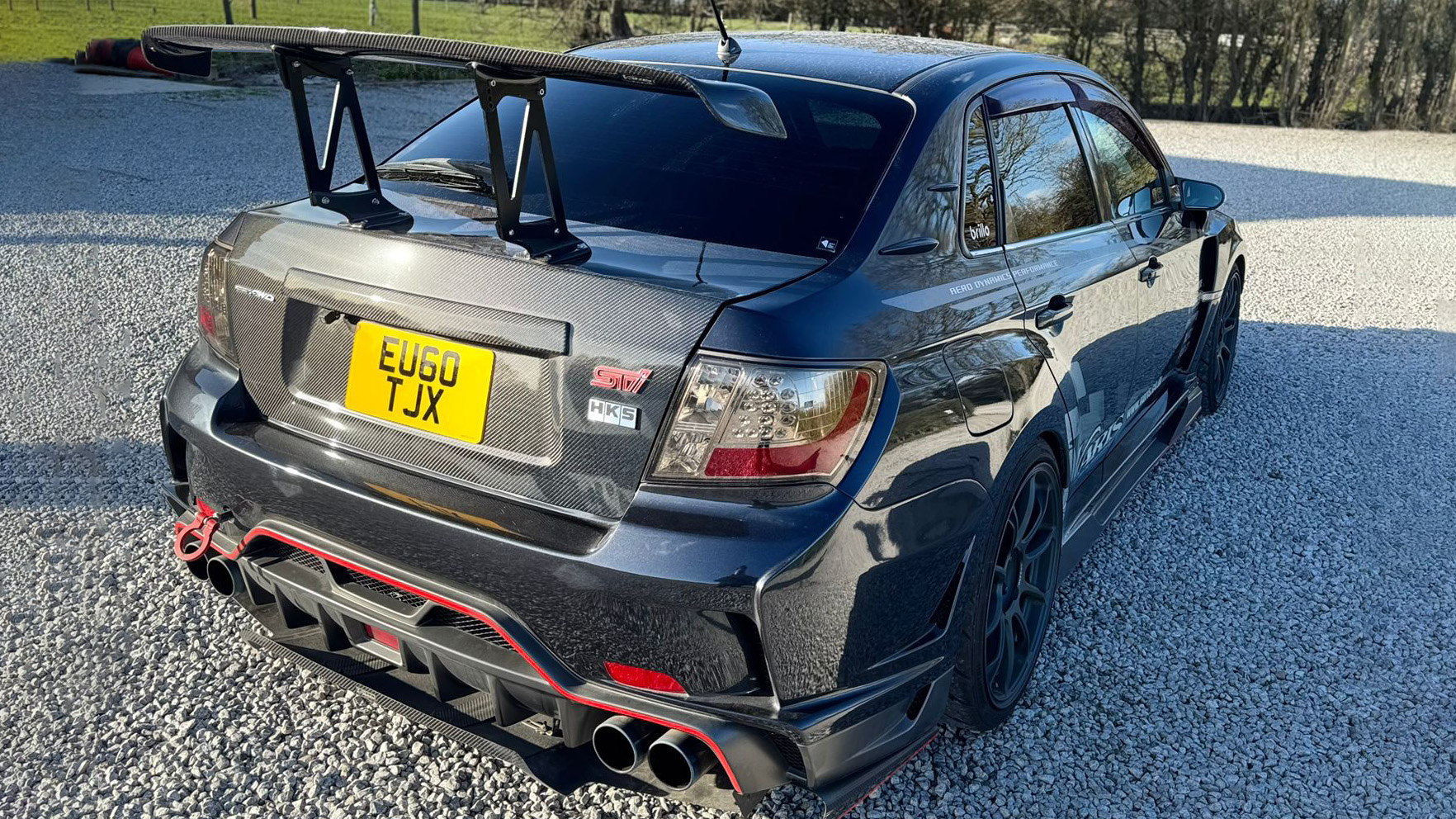 Rear three-quarter view of black Subaru Impreza showing large rear wing, quad exhausts and sporty diffuser, parked outdoors on gravel.