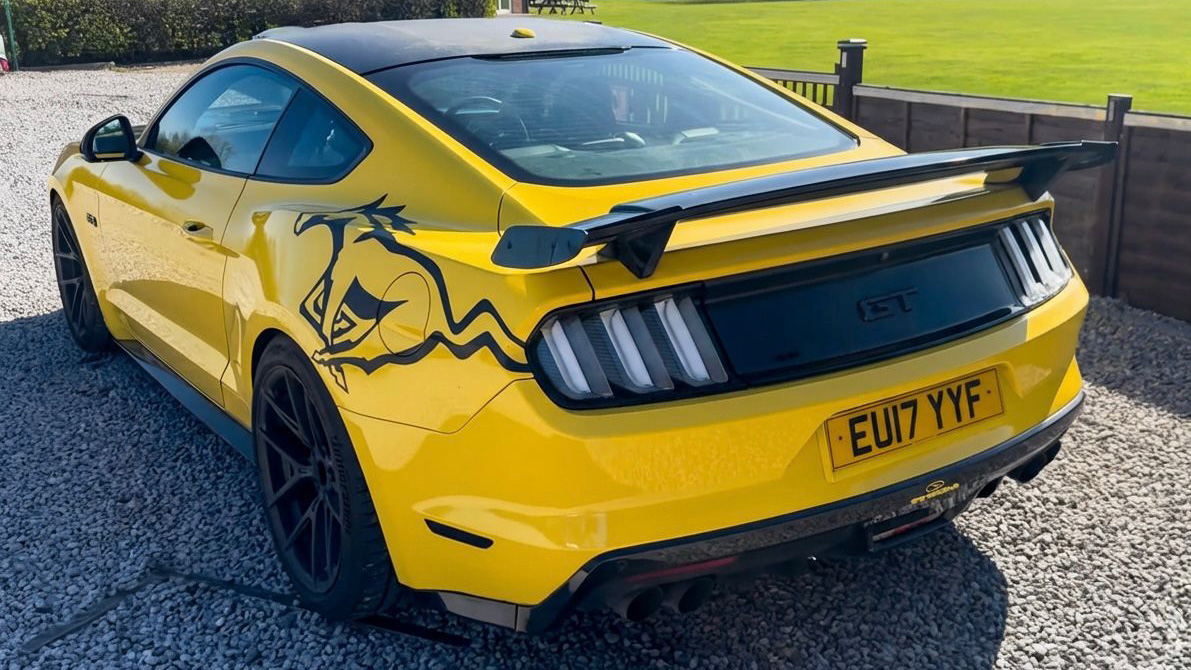 Rear view of yellow Ford Mustang GT with black spoiler and signature triple tail lights, parked outdoors on gravel.