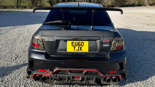 Direct rear view of black Subaru Impreza with large rear spoiler, quad exhaust system and UK number plate, parked on gravel with open landscape.