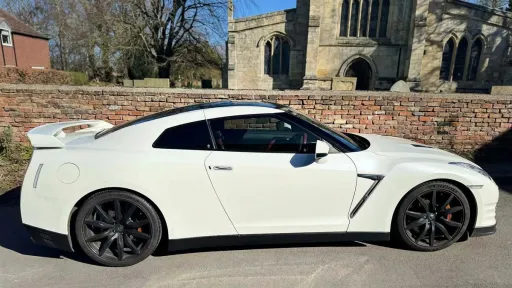 White Nissan GTR R35 parked near church grounds, side profile with black wheels, set against historic stone building backdrop.