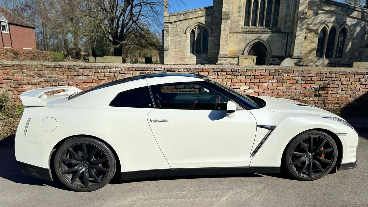 White Nissan GTR R35 parked near church grounds, side profile with black wheels, set against historic stone building backdrop.