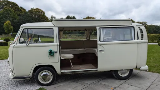 Old English White VW campervan front three-quarter view with cream interior, side door open, wedding ribbons on front, parked at countryside venue in Derbyshire