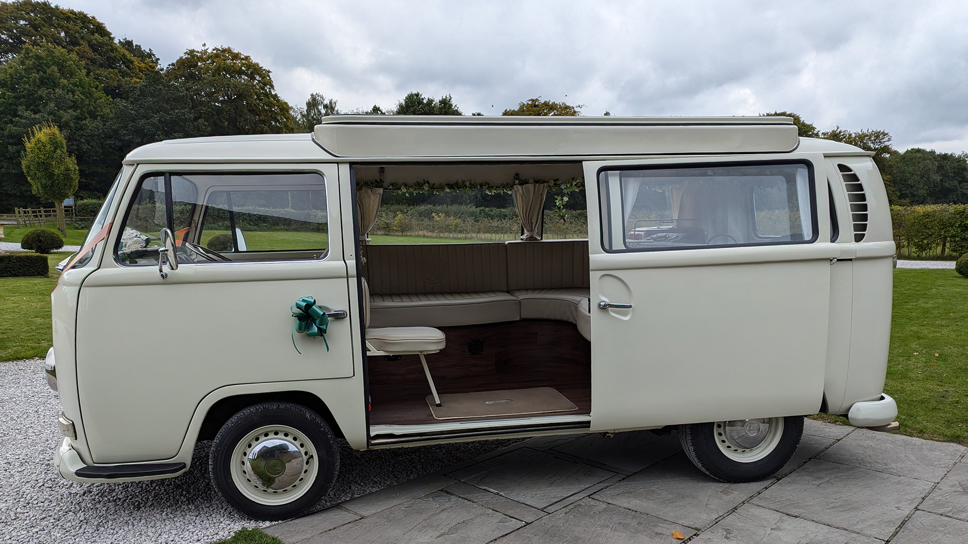 Old English White VW campervan front three-quarter view with cream interior, side door open, wedding ribbons on front, parked at countryside venue in Derbyshire