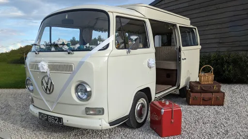 Old English White campervan front angle with period suitcases beside open side door, cream interior visible, parked outdoors