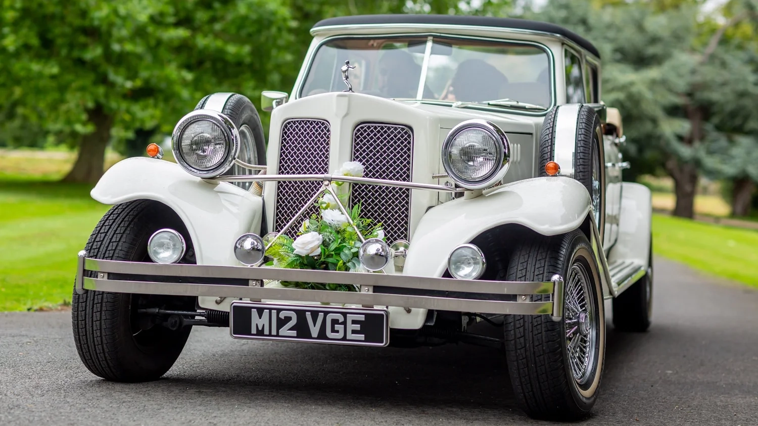 Front view white Beauford Landaulette wedding car with floral bonnet decoration parked on driveway