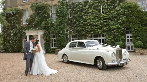 Bride and groom standing beside 1959 Rolls-Royce Silver Cloud in Old English White outside ivy covered venue