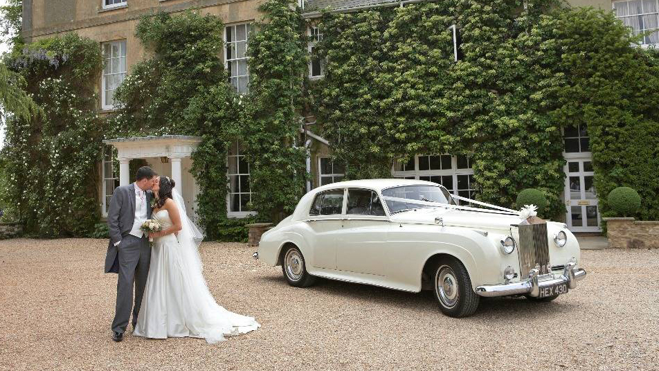 Bride and groom standing beside 1959 Rolls-Royce Silver Cloud in Old English White outside ivy covered venue