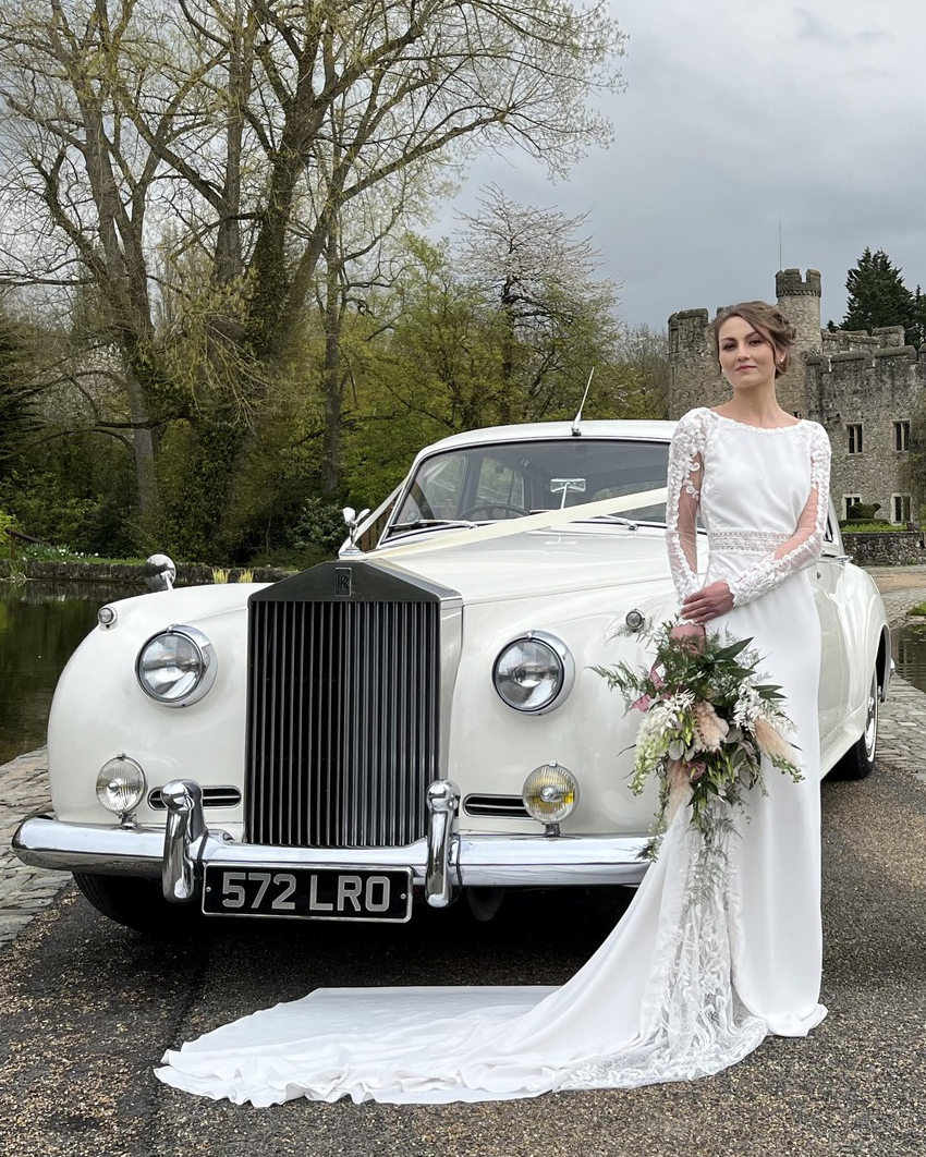 Bride holding a bouquet of flower and wearing a white wedding dress standing in front of a classic Rolls-Royce Silver Cloud at a Northumberland wedding venue