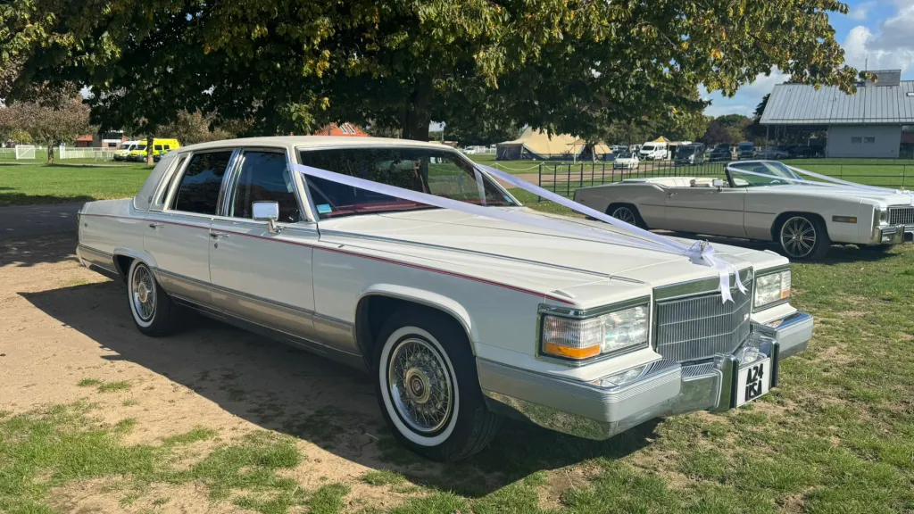 White 1990 Cadillac Brougham, front 3/4 view, chrome trim, whitewall tyres, tree-lined green backdrop. Wedding car hire East Anglia.