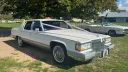 White 1990 Cadillac Brougham, front 3/4 view, chrome trim, whitewall tyres, tree-lined green backdrop. Wedding car hire East Anglia.