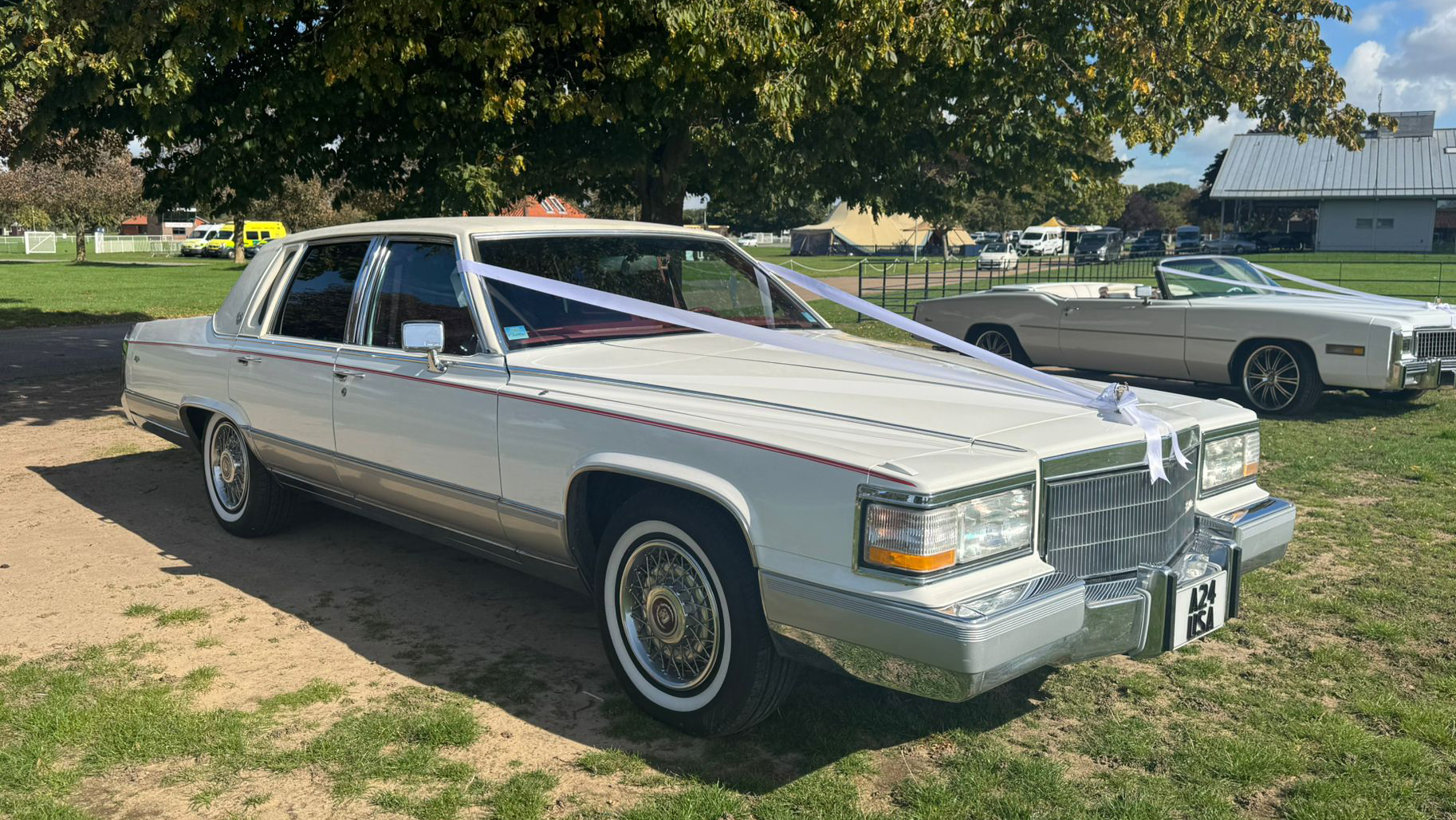 White 1990 Cadillac Brougham, front 3/4 view, chrome trim, whitewall tyres, tree-lined green backdrop. Wedding car hire East Anglia.
