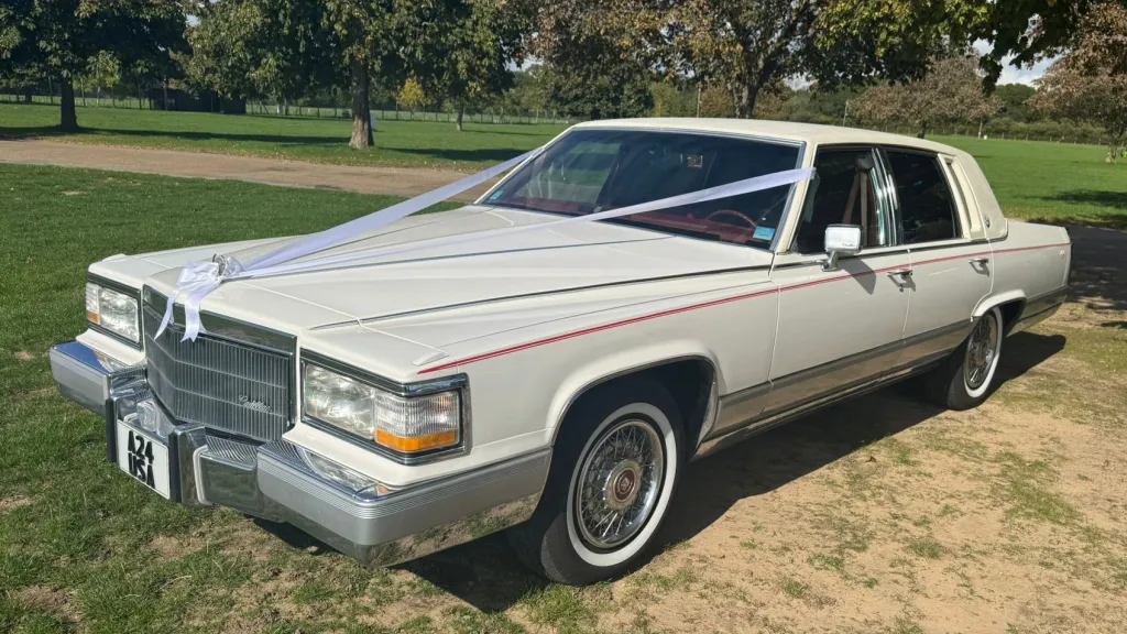 White 1990 Cadillac Brougham, left side view, whitewall tyres, chrome details, open parkland setting. Chauffeur-driven wedding car Suffolk.