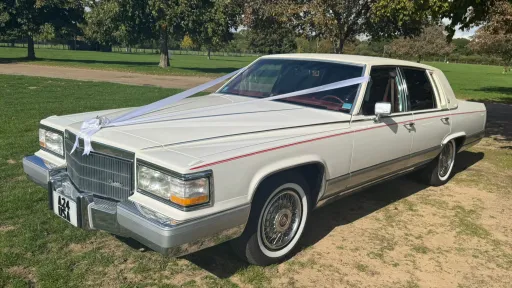 White 1990 Cadillac Brougham, left side view, whitewall tyres, chrome details, open parkland setting. Chauffeur-driven wedding car Suffolk.