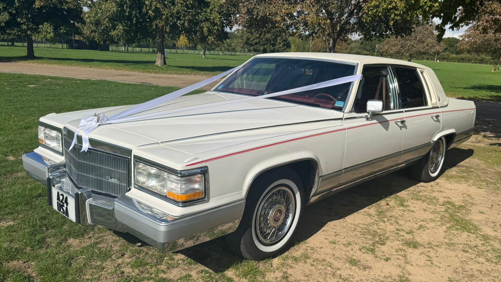 White 1990 Cadillac Brougham, left side view, whitewall tyres, chrome details, open parkland setting. Chauffeur-driven wedding car Suffolk.