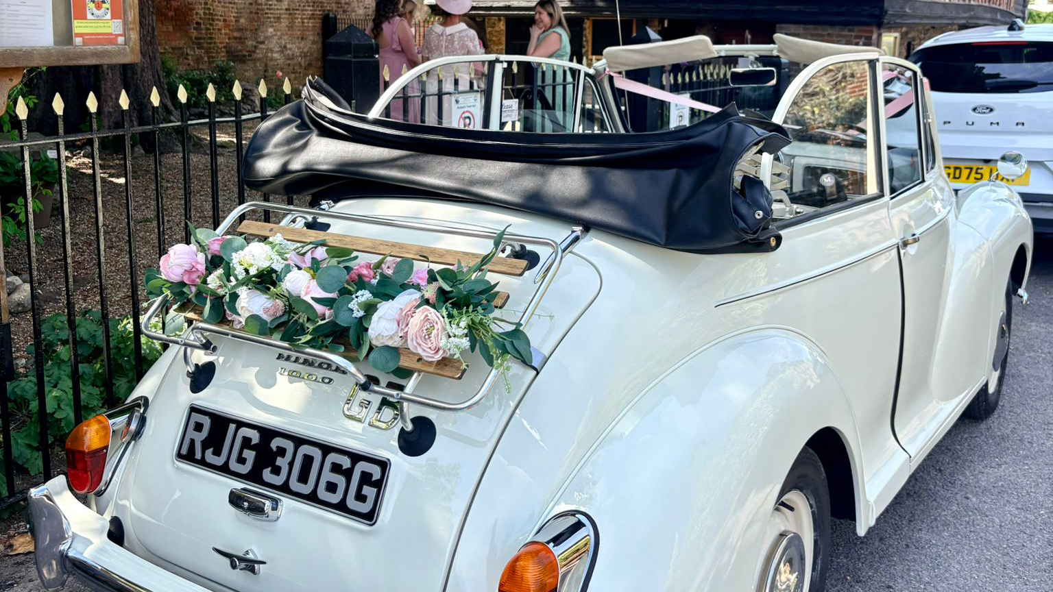 Bride and groom seated inside a 1969 Morris Minor Convertible in Old English White with black folded roof and pink wedding ribbons, photographed at a countryside wedding venue in Kent.