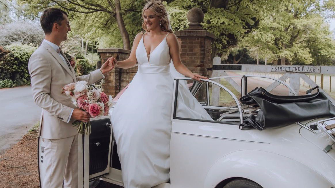 Bride stepping into a cream 1969 Morris Minor Convertible wedding car with chauffeur holding flowers beside the open door at a rustic countryside venue.