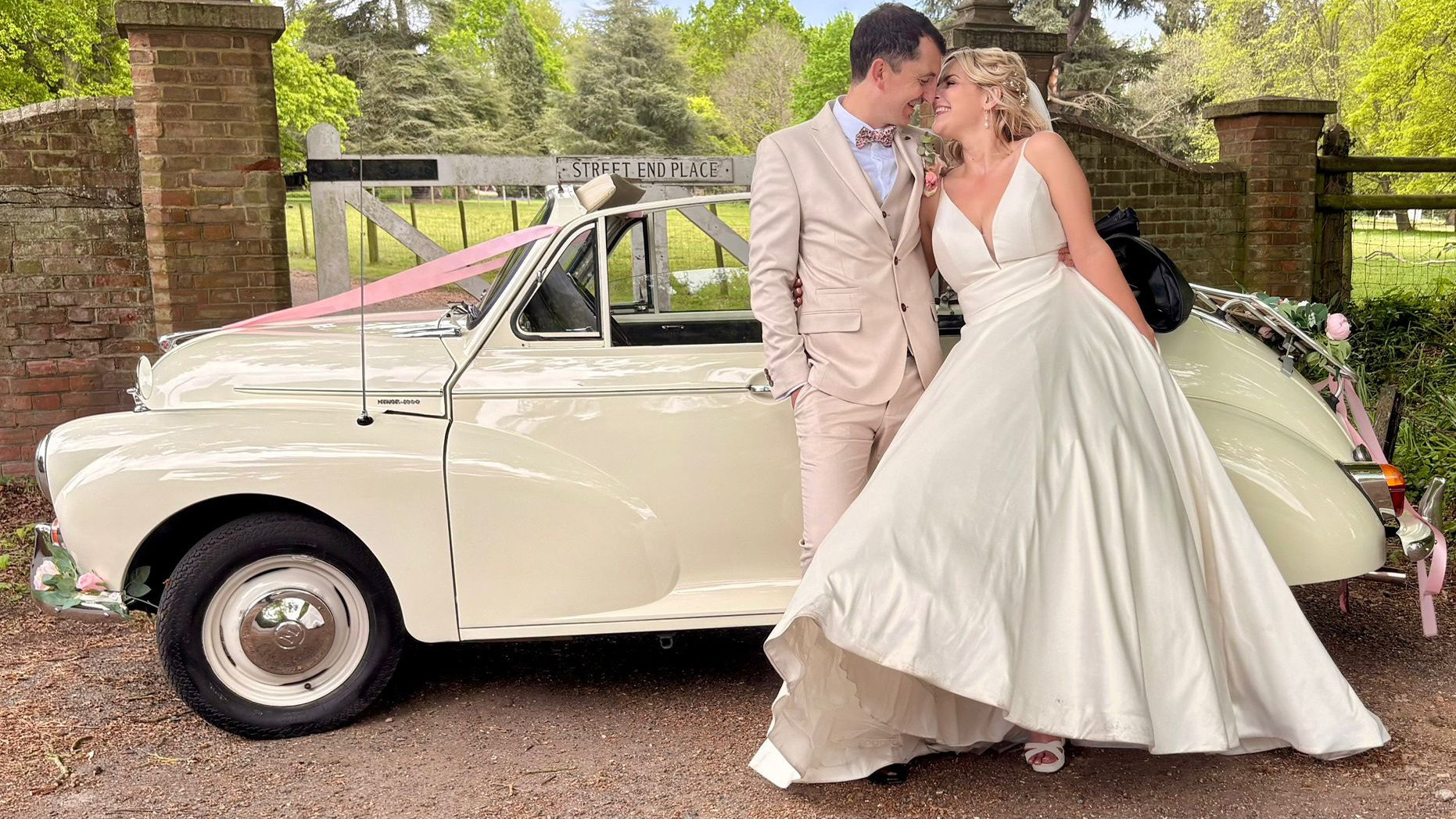 Bride and groom posing beside a 1969 Morris Minor Convertible in Old English White with black soft top roof folded down and chrome detailing.