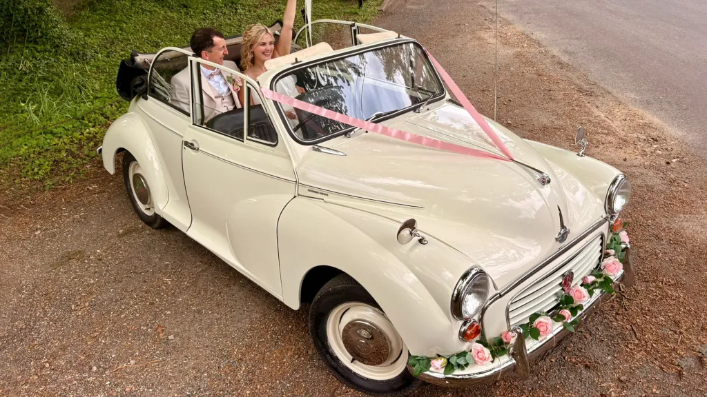 Front side view of a 1969 Morris Minor Convertible wedding car in Old English White with pink ribbons and floral bonnet decoration in Kent.