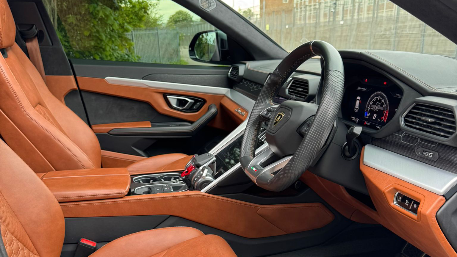 Driver side interior of Lamborghini Urus showing tan orange leather sports seats, digital dashboard and luxury steering wheel