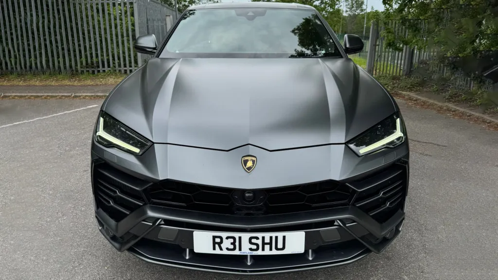 Front view of a matte grey Lamborghini Urus with black grille and black wheels parked on a quiet road surrounded by greenery