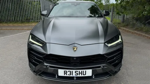 Front view of a matte grey Lamborghini Urus with black grille and black wheels parked on a quiet road surrounded by greenery