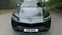 Front view of a matte grey Lamborghini Urus with black grille and black wheels parked on a quiet road surrounded by greenery