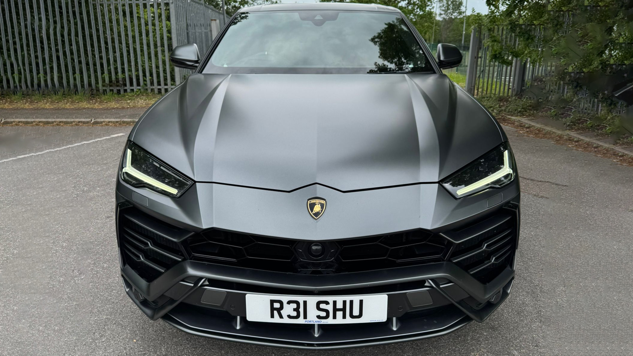 Front view of a matte grey Lamborghini Urus with black grille and black wheels parked on a quiet road surrounded by greenery