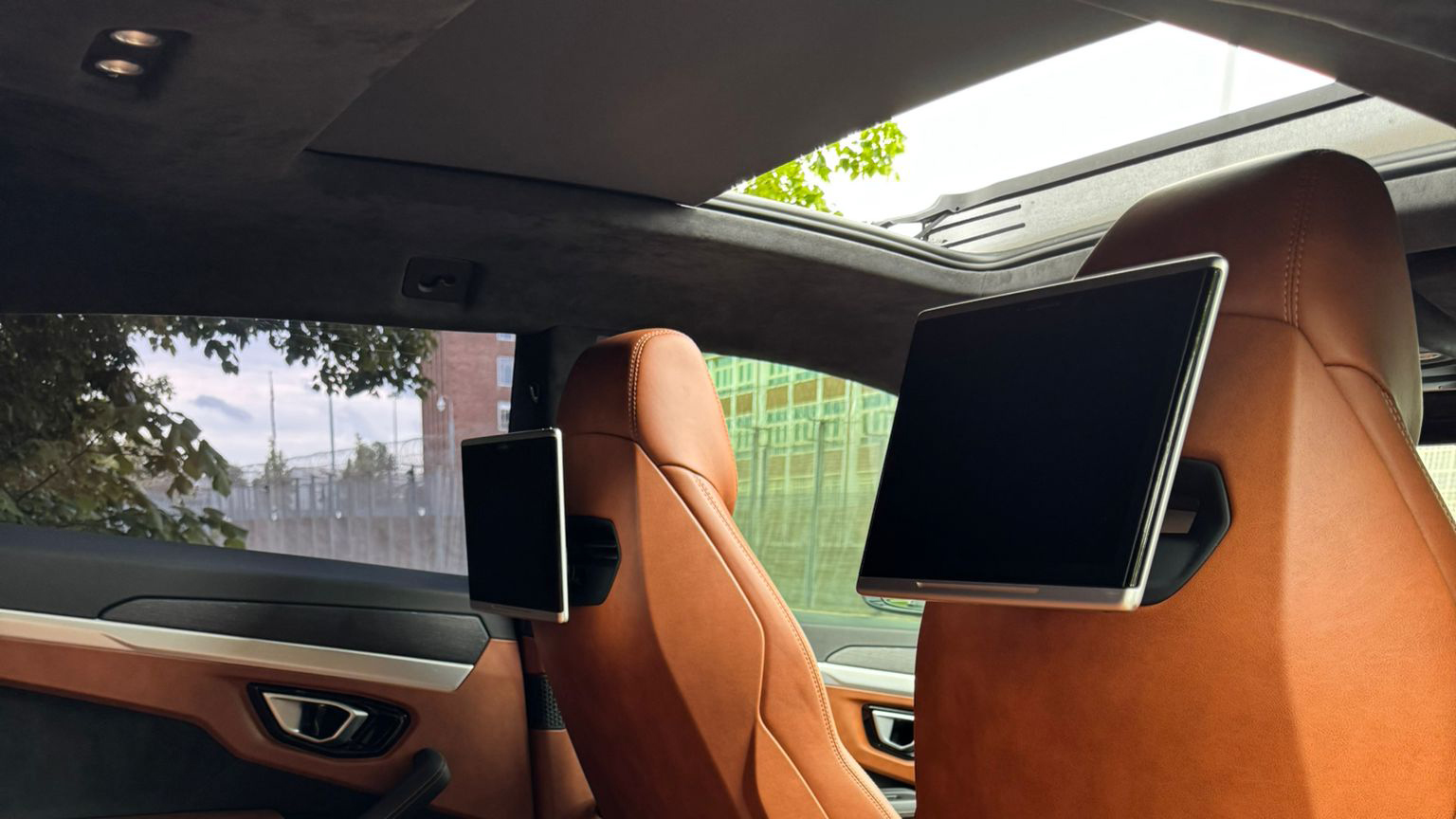 Luxury Lamborghini Urus interior with tan orange leather seats, panoramic sunroof and rear entertainment screens visible from the back cabin