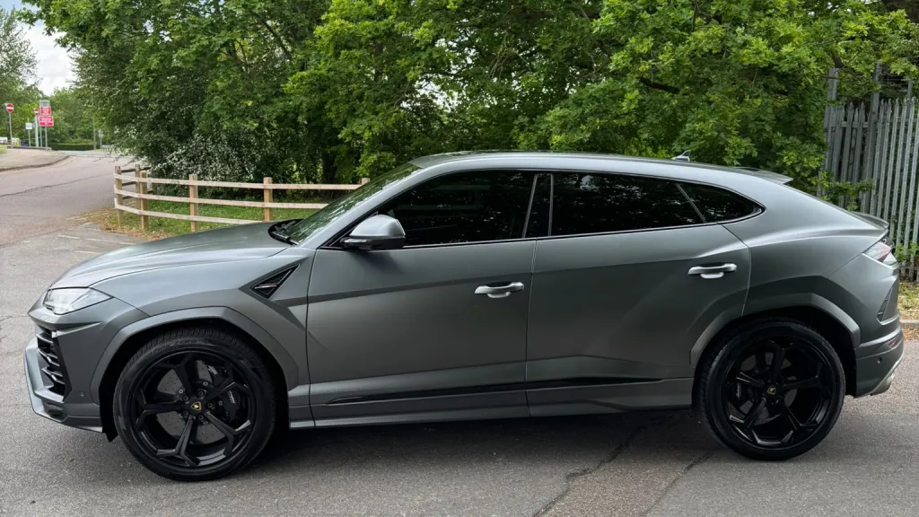Left side profile of matte grey Lamborghini Urus with black alloy wheels parked beside trees and fencing