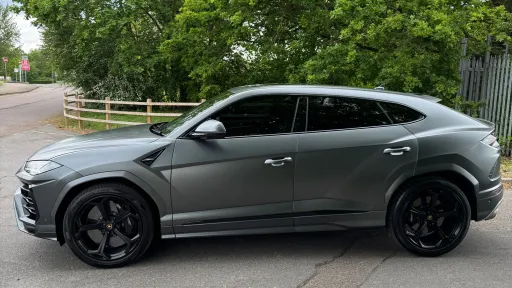 Left side profile of matte grey Lamborghini Urus with black alloy wheels parked beside trees and fencing