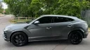 Left side profile of matte grey Lamborghini Urus with black alloy wheels parked beside trees and fencing