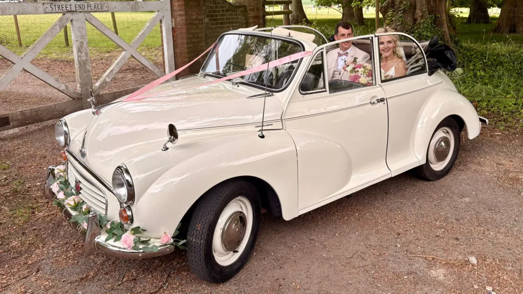 1969 Morris Minor Convertible wedding car driving through a countryside venue in Kent with bride and groom smiling from the rear seats.