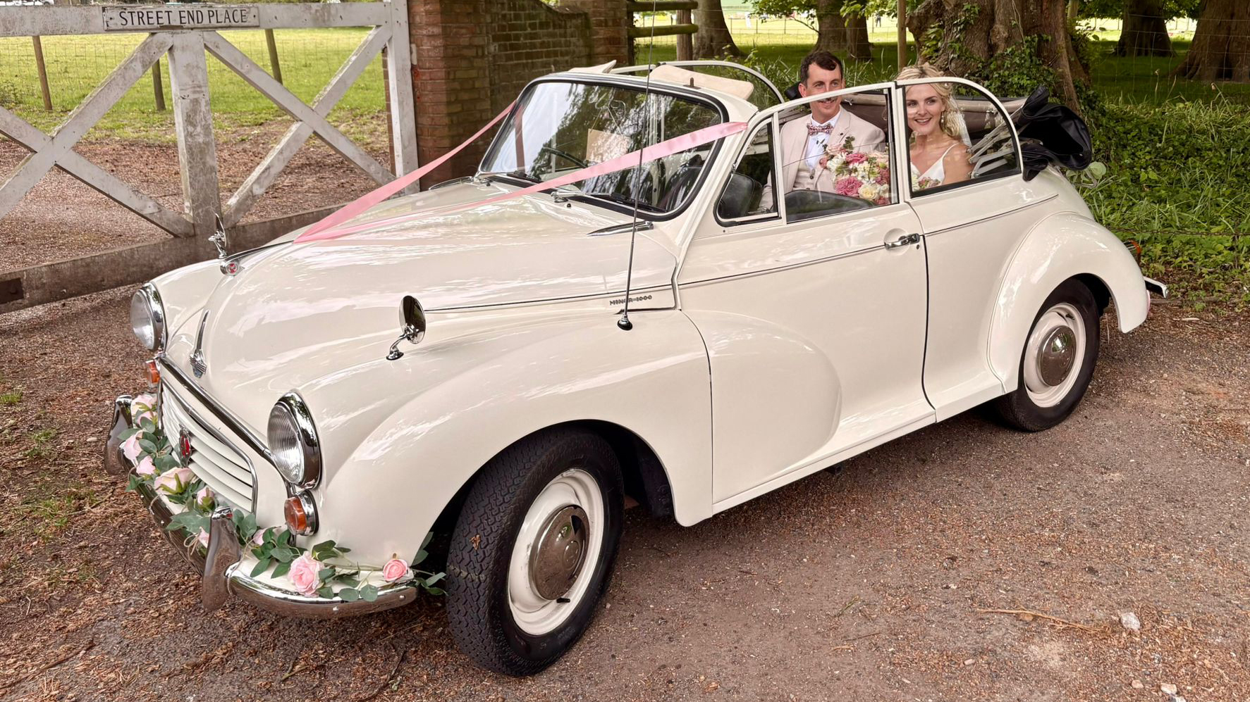 1969 Morris Minor Convertible wedding car driving through a countryside venue in Kent with bride and groom smiling from the rear seats.