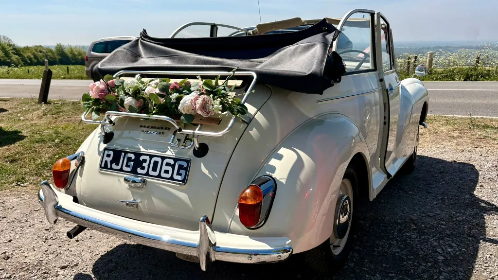 Rear view of an Old English White Morris Minor Convertible wedding car with folded black roof and pastel flower arrangement attached to the rear rack.