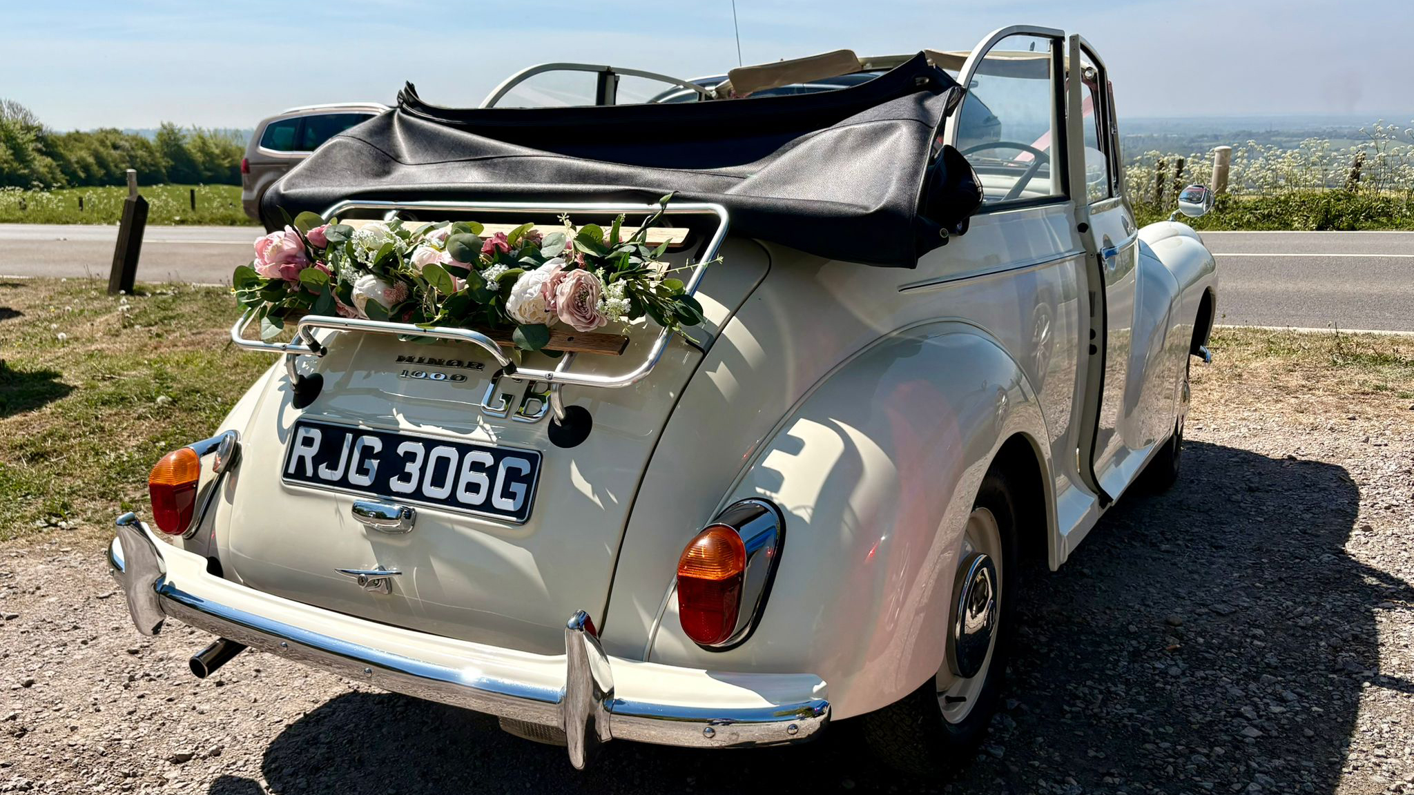 Rear view of an Old English White Morris Minor Convertible wedding car with folded black roof and pastel flower arrangement attached to the rear rack.