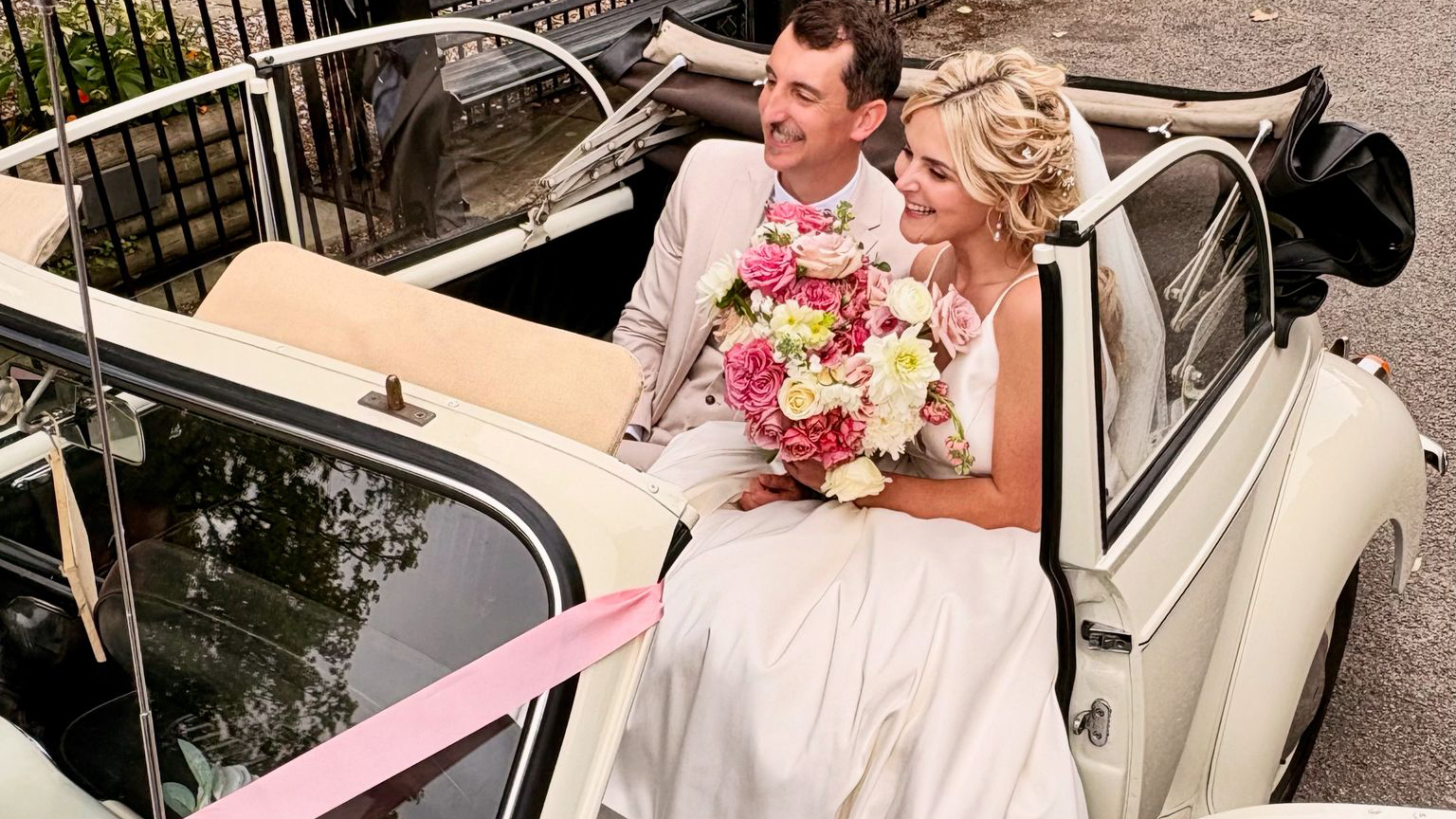 Happy couple seated inside a 1969 Morris Minor Convertible in Old English White with pink wedding ribbons and chrome windscreen surround.