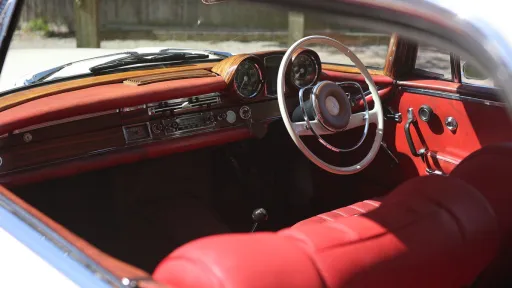 Driver’s view inside Mercedes 250 SE Coupe showing red leather dashboard, wood trim and classic steering wheel.
