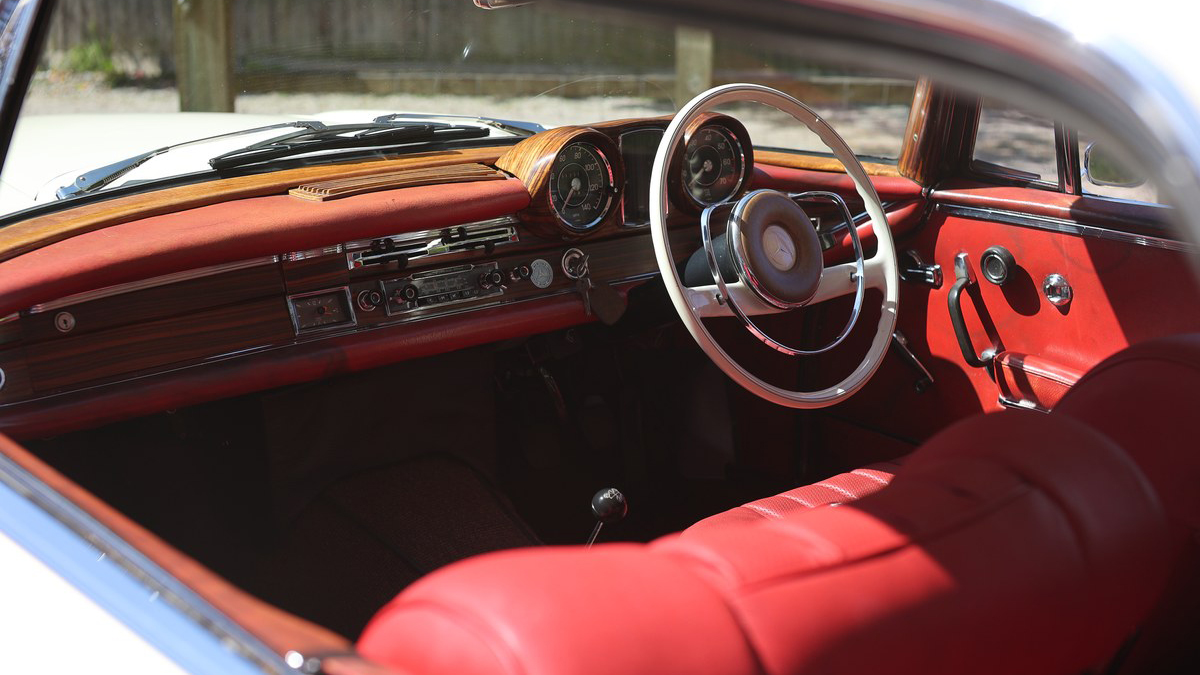 Driver’s view inside Mercedes 250 SE Coupe showing red leather dashboard, wood trim and classic steering wheel.