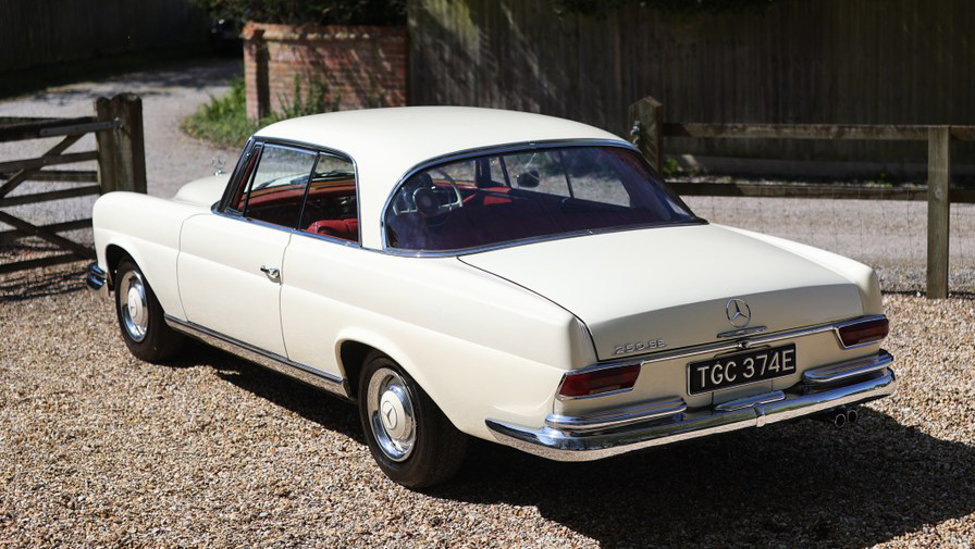 Rear view of ivory Mercedes 250 SE Coupe wedding car parked on gravel with red interior visible.