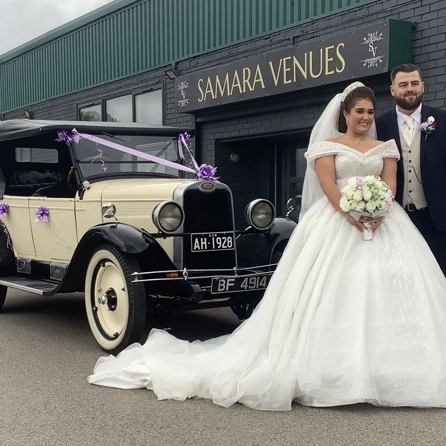 Bride and groom posing outside Samara Venues in Ashford, Kent beside a vintage cream and black wedding car decorated with purple ribbons and bows, with the bride holding a white bouquet in front of the venue entrance.