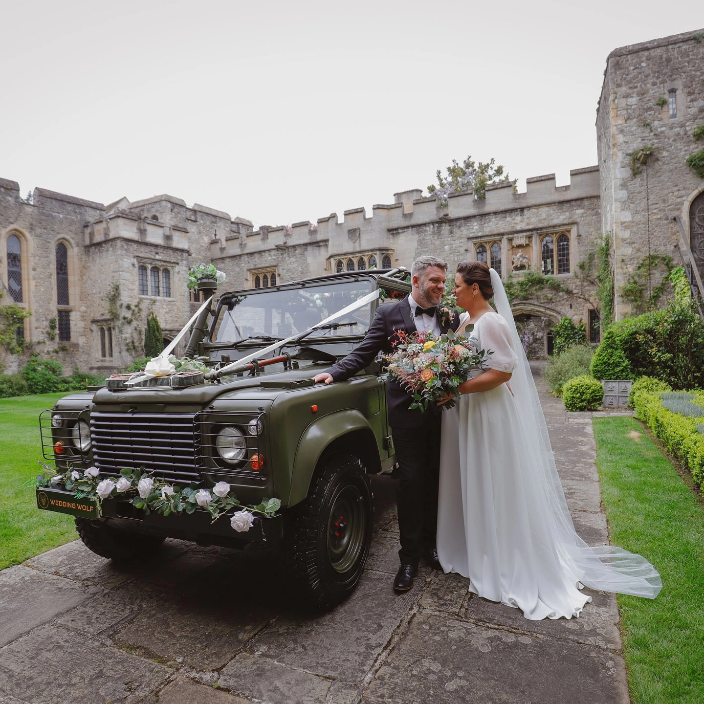 Bride and groom standing beside a classic green Land Rover wedding car at Allington Castle in Maidstone, Kent, with the bride holding a colourful bouquet against the historic castle backdrop.