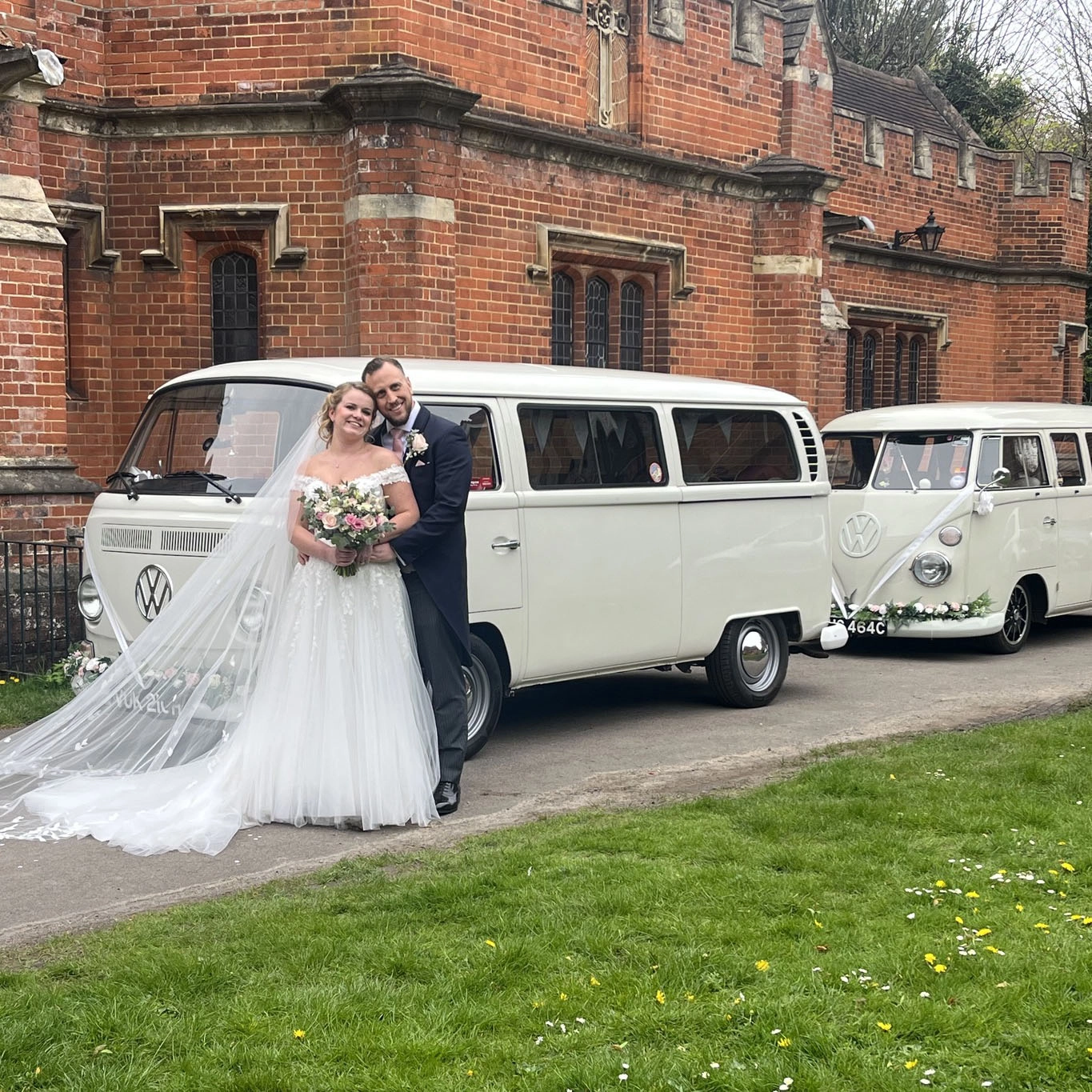 Bride and groom posing beside two classic white VW campervan wedding vehicles at Reach Court Farm in Dover, Kent, with the bride’s long veil flowing across the grass outside the red brick venue.