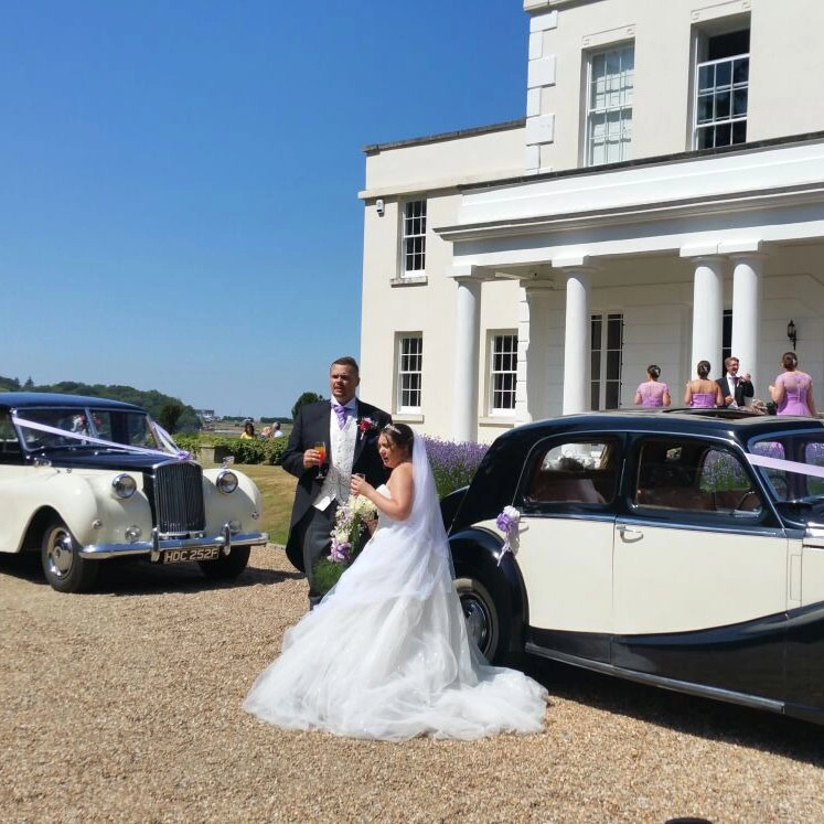 Bride and groom beside two classic wedding cars at Little Hermitage in Rochester, Kent, featuring a black and cream vintage car and Rolls-Royce under bright summer skies.